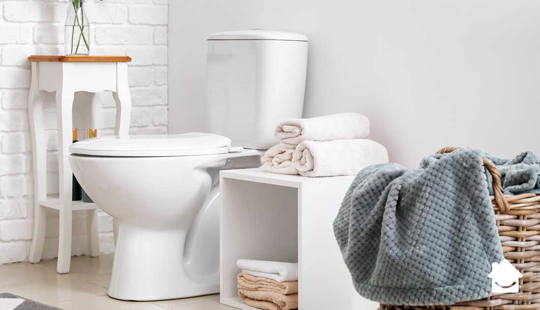 An image of a white bathroom, including a toilet, a white unit containing towels, and a woven laundry basket in the foreground.