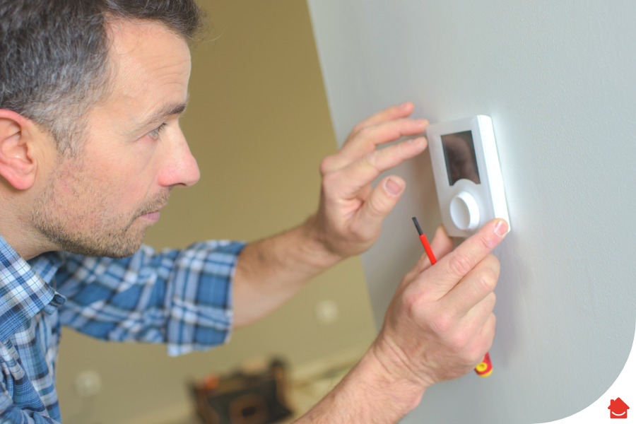 An image of a man in a blue plaid shirt holding a red screwdriver. He is adjusting a thermostat.