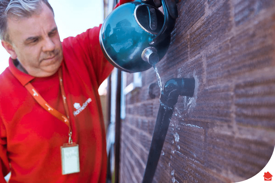 A HomeServe engineer pouring boiling water from a kettle over a boiler condensate pipe outside.