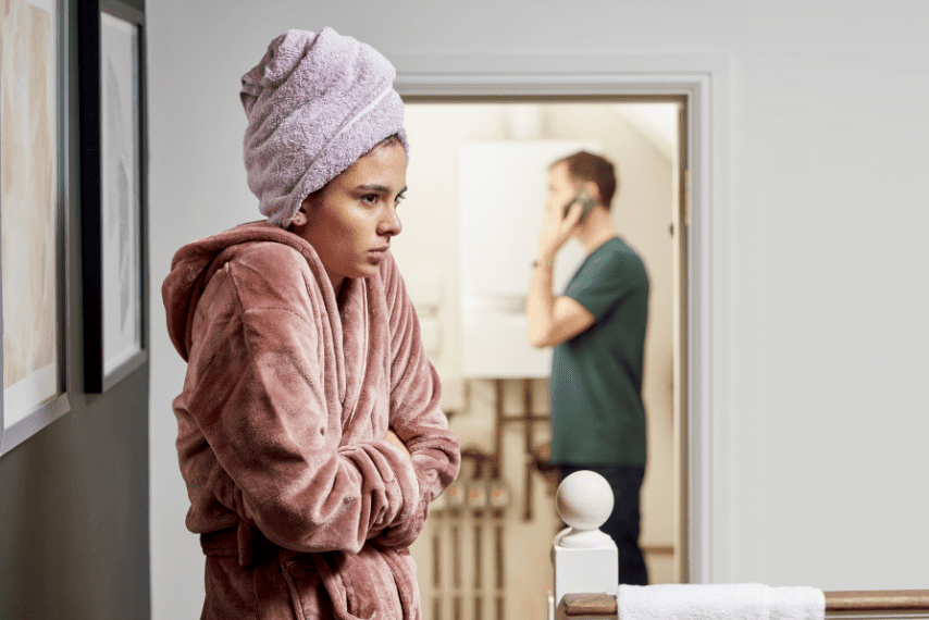 A girl in a hallway shivering, wearing a dressing gown and a towel on her head. Her dad is on the phone in the background.